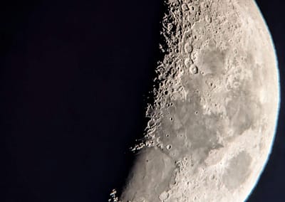 A zoomed in crescent moon with large craters along the line separating the light and dark parts of the moon.